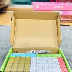 The box opened on a grey floor, the brown carton lid folded back to show a green tray packed with blocks sorted into colour bands.