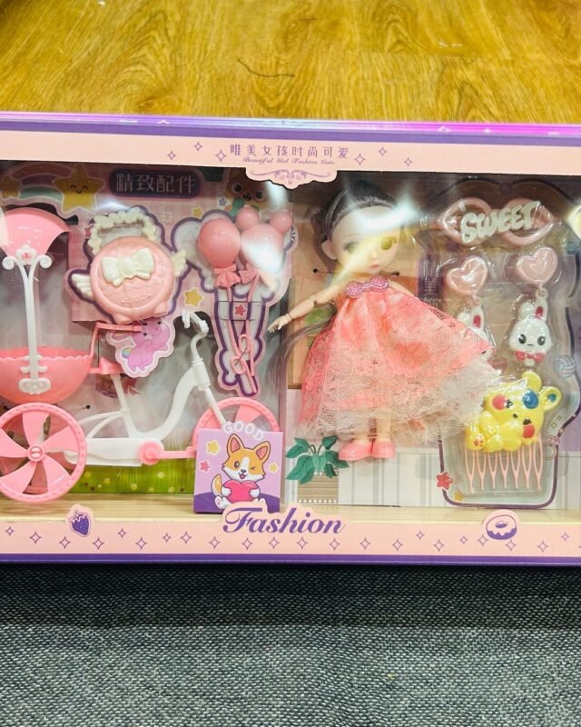 The long lilac box on a grey floor: a doll in a peach dress stands beside a pink tricycle, with balloons, a basket and a small rabbit around her.