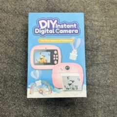 The sealed blue retail box standing on grey fabric, printed with the pink camera and a black-and-white photograph sliding out of its slot.