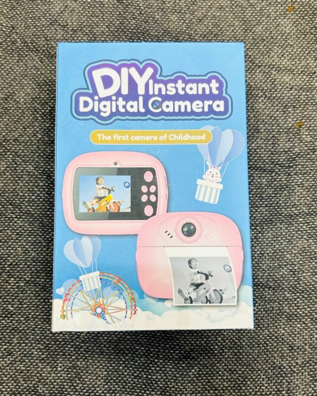 The sealed blue retail box standing on grey fabric, printed with the pink camera and a black-and-white photograph sliding out of its slot.