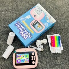The contents seen from above with the box lying at the top of the frame, the pink camera below it beside a paper roll, the cable and the sealed pack of pens.