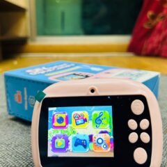 Close on the pink camera indoors, its colour screen showing nine app icons, the box out of focus behind it.